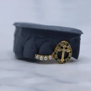 Custom gold two-piece grillz: an oval cross cap set with an emerald and diamond border, paired with a pearl-set connector band, shown on a dental mold
