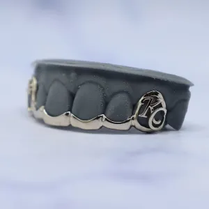 Custom white gold bottom grillz with engraved script initials on the right canine cap, shown fitted on a dental cast from the side